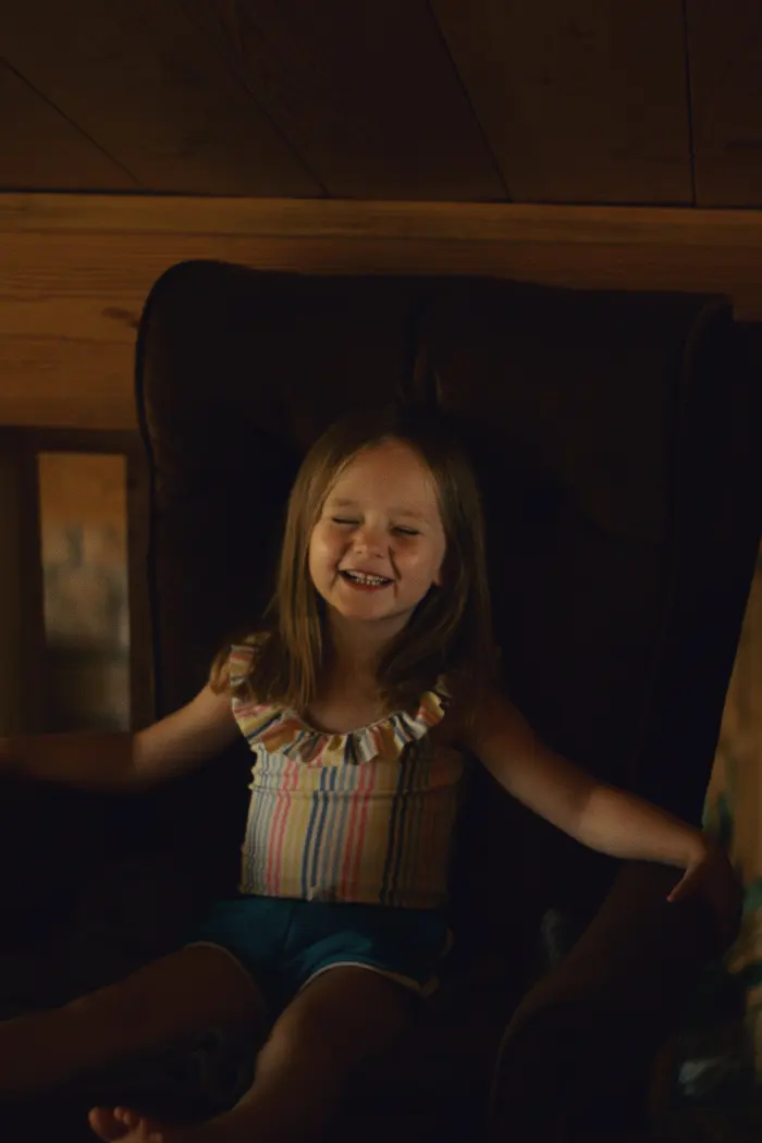 A little girl laughs with her eyes shut in a big armchair inside a wooden cabin