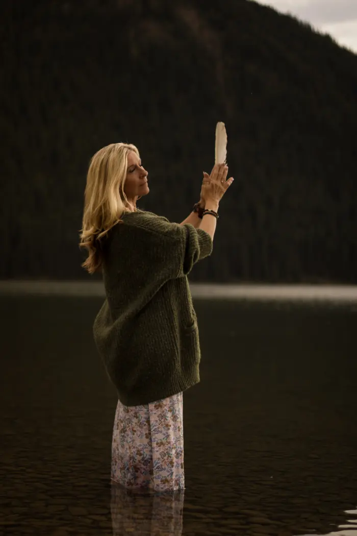 A woman stands in a lake holding a white feather up in front of a dark forested mountain