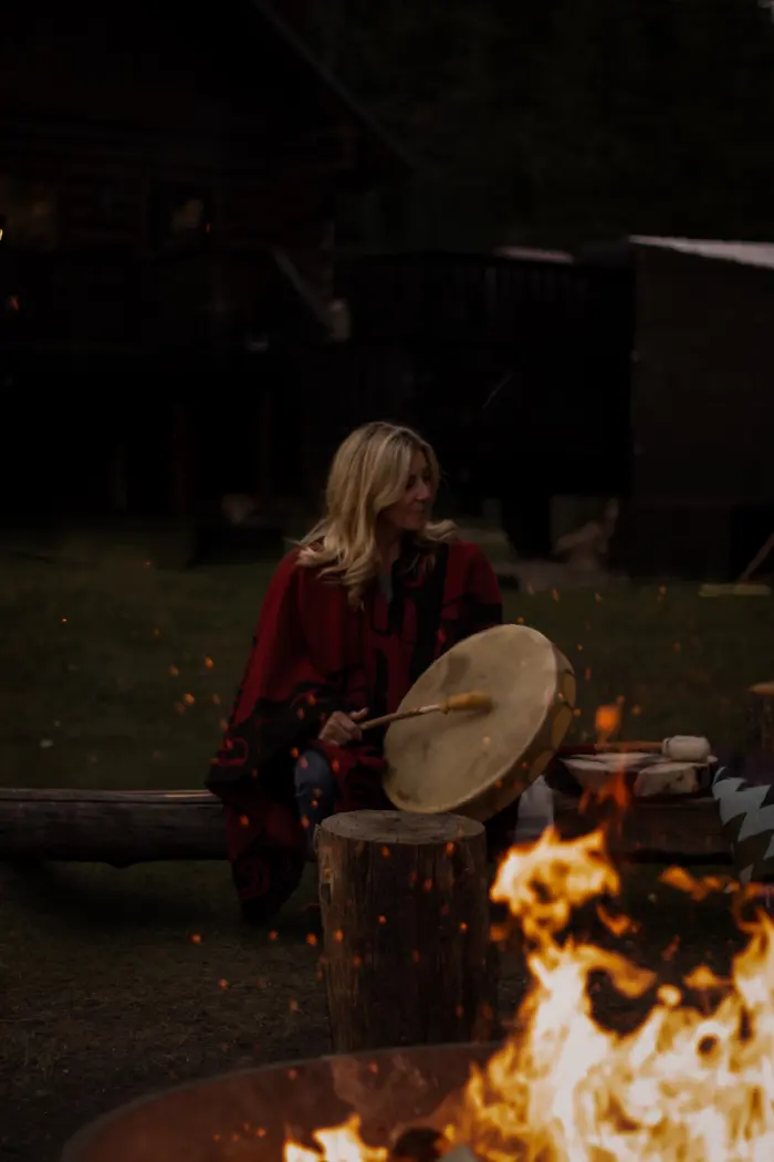 A woman wrapped in a red blanket plays a hand drum beside a crackling fire