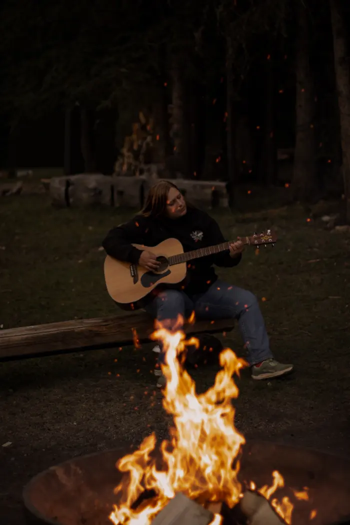A woman plays an acoustic guitar beside a campfire as sparks drift up into the trees