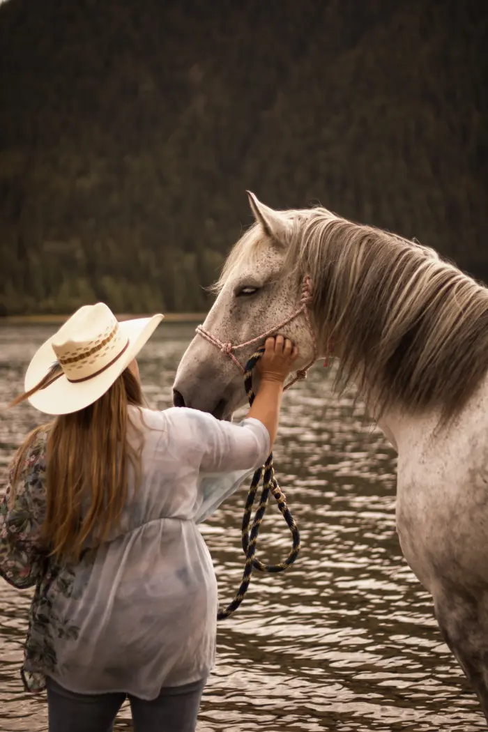 A woman in a straw cowboy hat rests her hand on a grey horse standing in a lake