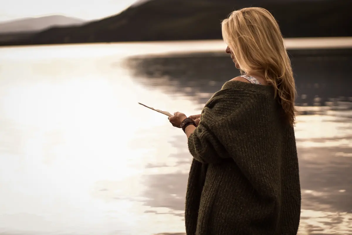 A woman in a moss green cardigan holds a feather at the edge of a shimmering lake