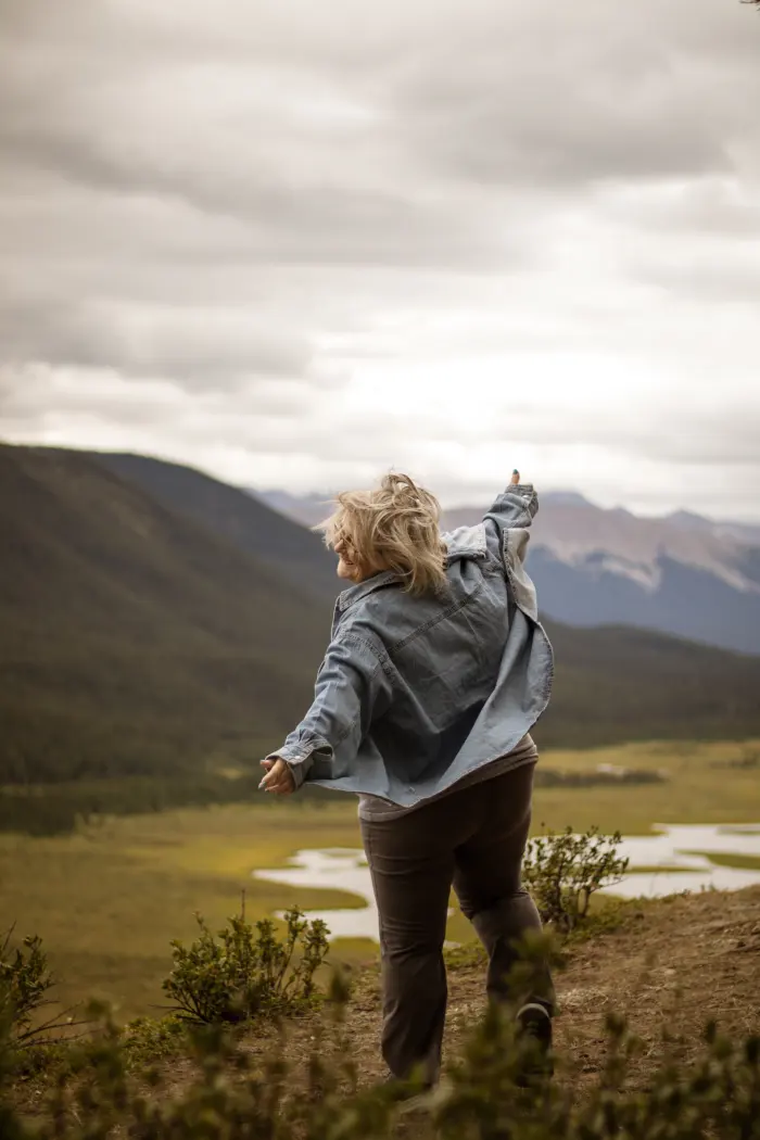 A woman in a denim jacket throws her arms out on a ridge above a wide mountain valley