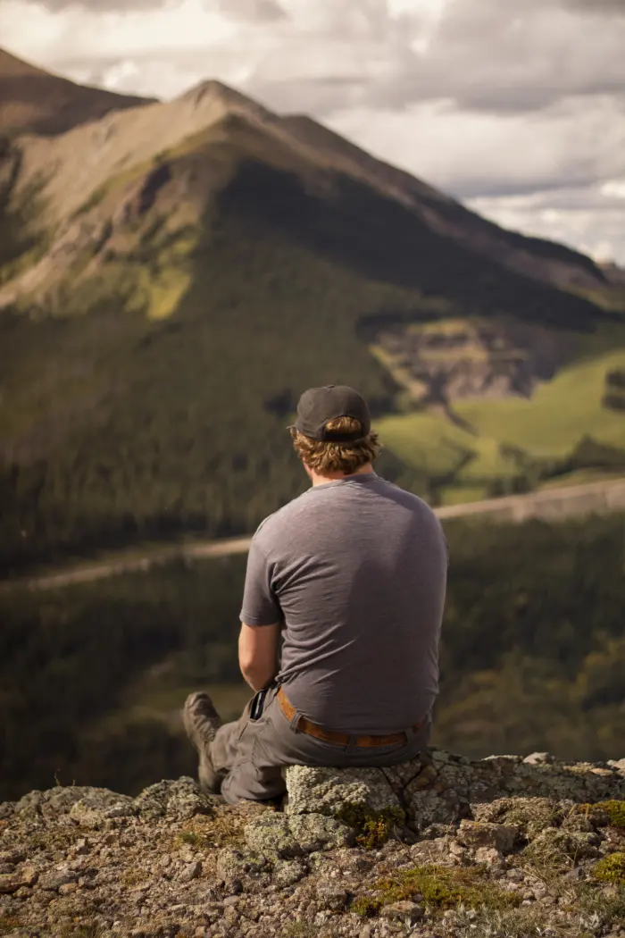 A young man sits on a lichen covered rock looking out over a mountain valley